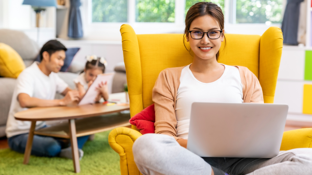 a smiling virtual assistant woman sitting on a yellow accent couch with laptop on her lap and at the background are a man playing some toys with a child depicting a positive vibe despite of challenges as a virtual assistant